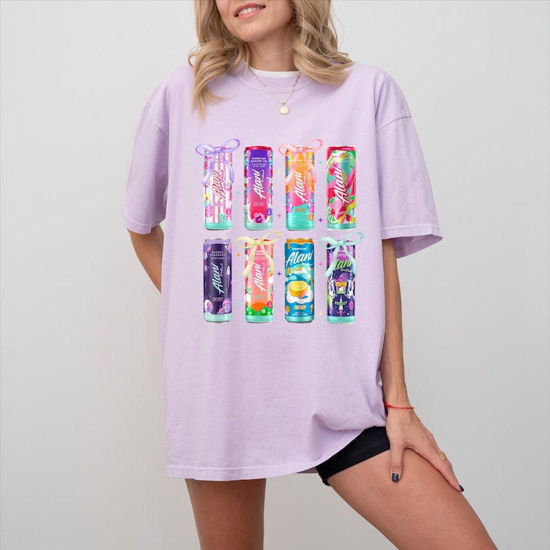 May include: A light purple t-shirt featuring a graphic of nine beverage cans with the brand name "Alan" on them. The cans are in various colors and designs, with decorative bows. The shirt is worn by a person.