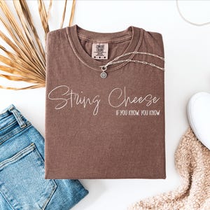 May include: A brown t-shirt with the words "String Cheese" and "If you know, you know" printed on the front. The shirt is paired with blue jeans, white sneakers, and a silver necklace. A dried palm leaf adds a natural element to the composition.