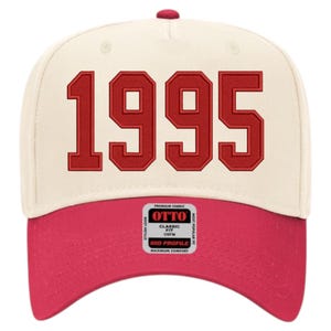 May include: A baseball cap with a cream-colored crown and a red bill. The number "1995" is embroidered in red on the front. The cap has a mid-profile design and features a "OTTO" brand label.