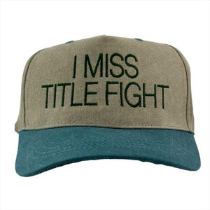 May include: A baseball cap with a khaki crown and a teal blue brim. The cap features the embroidered text "I MISS TITLE FIGHT" in dark green thread. The hat is a classic six-panel design.