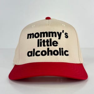 May include: A baseball cap with a cream-colored crown and a red brim. The text "mommy's little alcoholic" is embroidered in black on the front of the cap. The cap has a red button on top.