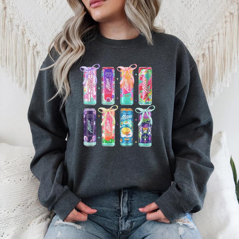 Puede incluir: Sudadera gris oscuro con un gr&aacute;fico colorido de nueve latas de bebidas. Cada lata tiene un dise&ntilde;o y sabor diferente, con la marca "Alani" visible. Las latas est&aacute;n decoradas con lazos.