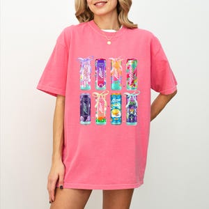 May include: A pink t-shirt featuring a collection of colorful beverage cans. Each can displays the brand name "Alan" and is adorned with a decorative bow. The cans showcase various fruit-inspired designs and colors.