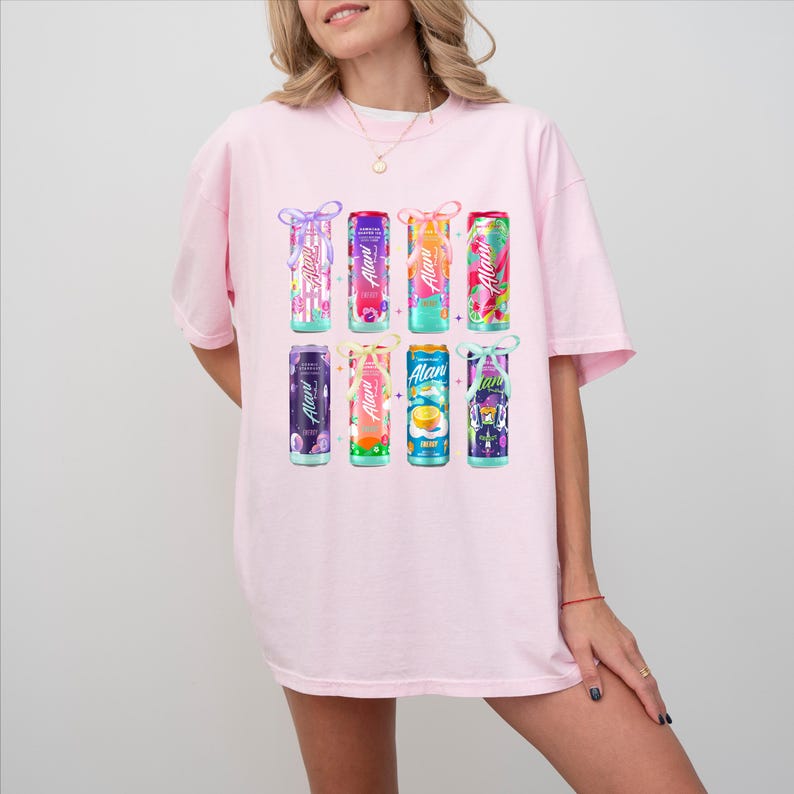 May include: A light pink t-shirt featuring a graphic of eight colorful beverage cans. Each can has a different design and flavor, with the brand name "Alani" in a cursive font. The cans are arranged in two rows of four.