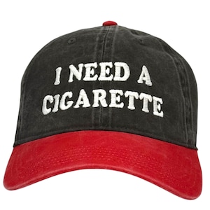 May include: A black baseball cap with a red brim and button. The phrase "I NEED A CIGARETTE" is embroidered in white across the front of the cap. The cap is made of a soft, worn material.