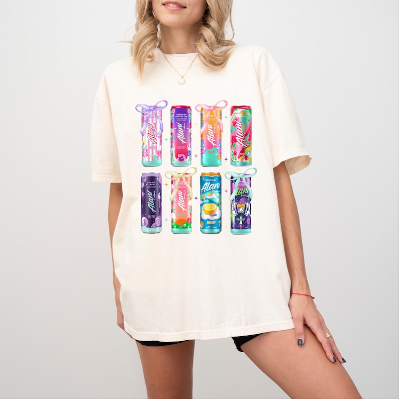May include: Cream-colored t-shirt featuring a colorful graphic of eight beverage cans. Each can has a different design and flavor name, with the brand name "Alani" prominently displayed. The cans are arranged in two rows of four.