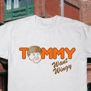Puede incluir: Camiseta blanca con un gráfico naranja de la cara de un hombre de dibujos animados y el texto "TOMMY Want Wingy".