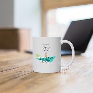 May include: White ceramic mug featuring a cartoon illustration of a man with glasses and the text "CLEVELAND ROCKS" in black and teal. The mug has a curved handle and is set on a wooden surface.