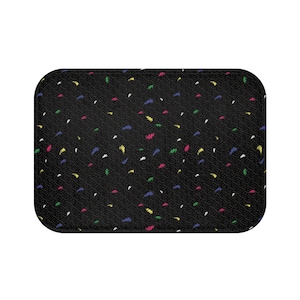 May include: Rectangular black bath mat with a pattern of small white dots and colorful, abstract brushstrokes in yellow, green, pink, blue, and white. The mat has rounded corners and a textured surface.