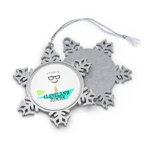 May include: A silver snowflake ornament with a cartoon image of a man wearing glasses and the text "CLEVELAND ROCKS". The ornament has a silver string for hanging.