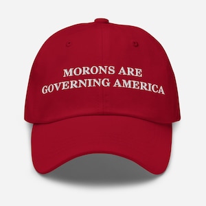 Könnte beinhalten: Rote Baseballkappe mit weiß gesticktem Text „MORONS ARE GOVERNING AMERICA“. Die Kappe hat einen gebogenen Schirm und eine abgerundete Krone. Der Hut ist aus einem weichen Material gefertigt.