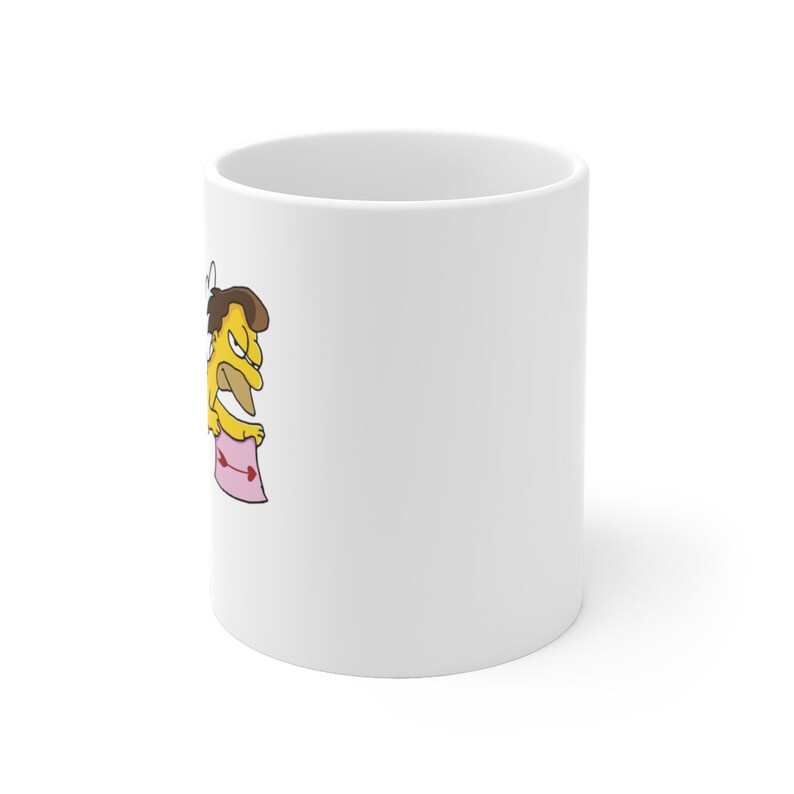 May include: White ceramic mug with a cartoon illustration of a yellow character with brown hair wearing a hat and holding a pink sign with a red dumbbell on it.