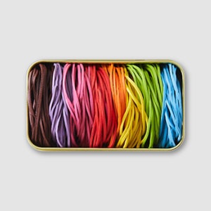 May include: A rectangular gold tin containing a selection of brightly coloured cords. The cords are arranged in a rainbow pattern, including brown, purple, pink, red, orange, yellow, green, and blue. The cords appear to be made of a flexible material.