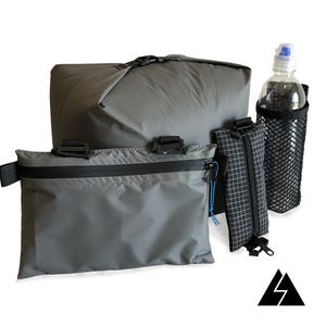 Può includere: Un set di quattro pochette grigie con cerniera, tra cui una grande pochette rettangolare, una pochette rettangolare più piccola, una piccola pochette quadrata e una pochette a rete con una bottiglia d'acqua in plastica trasparente all'interno.