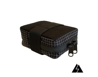May include: A black and white grid patterned zippered pouch with a black strap. The pouch is made of a durable fabric and has a zipper closure.