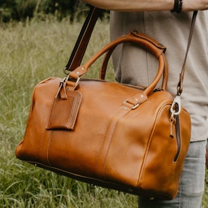 May include: A brown leather duffel bag with a shoulder strap and top handles. The bag has a rectangular shape with a zipper closure and a luggage tag. The bag is carried by a person in a field of tall grass.