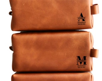 May include: Six brown leather toiletry bags stacked on top of each other. Each bag has a personalized monogram and the text "Groomsman" and the date "May 31, 2024".