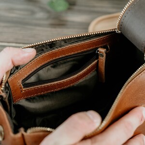 May include: A brown leather travel bag with a zipped closure and a smaller interior pocket. The bag is open and the interior is lined with a dark fabric.