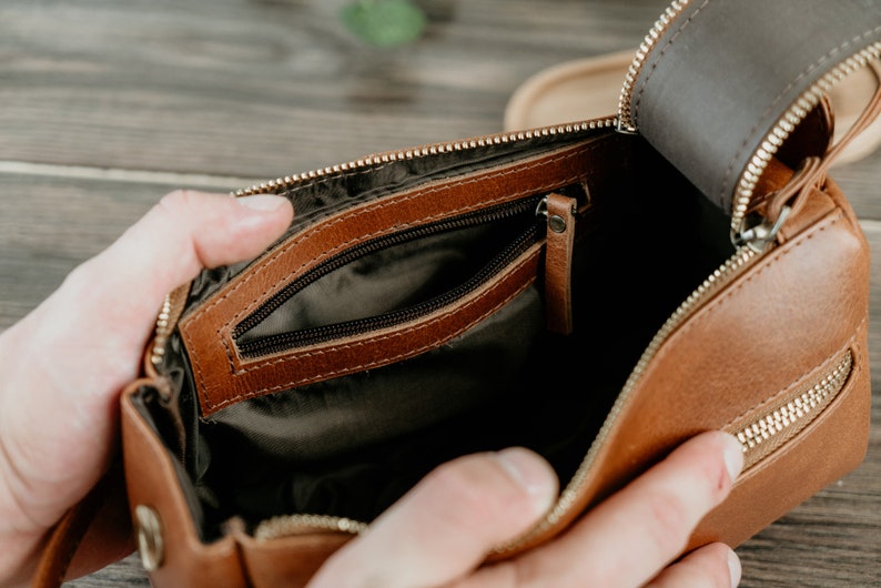 May include: A brown leather travel bag with a zipped closure and a smaller interior pocket. The bag is open and the interior is lined with a dark fabric.