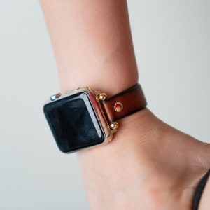 Op de afbeelding: Een zilveren Apple Watch met een bruin leren bandje en gouden accenten. Het horloge heeft een zwart display en het bandje heeft een rood accent.