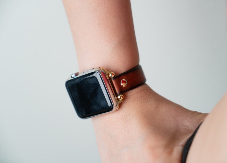 Op de afbeelding: Een zilveren Apple Watch met een bruin leren bandje en gouden accenten. Het horloge heeft een zwart display en het bandje heeft een rood accent.