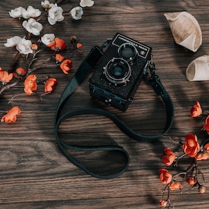 May include: A black vintage camera with a leather strap, surrounded by white and orange flowers on a wooden surface.