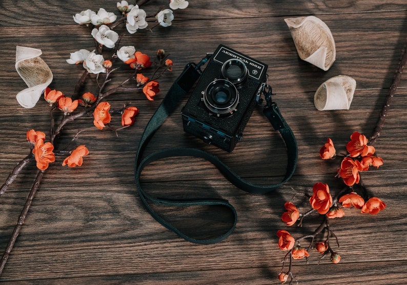 May include: A black vintage camera with a leather strap, surrounded by white and orange flowers on a wooden surface.