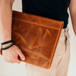 May include: A brown leather laptop sleeve with a flap closure. The sleeve is being held by a person wearing a black shirt and beige pants.