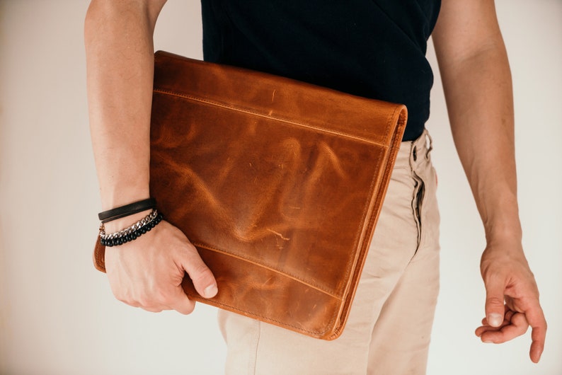 May include: A brown leather laptop sleeve with a flap closure. The sleeve is being held by a person wearing a black shirt and beige pants.