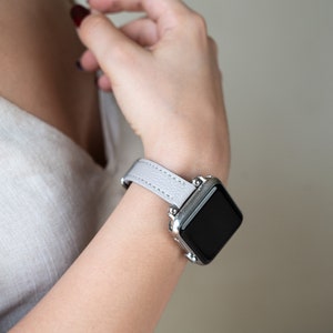 May include: A light gray leather watch band with a silver smartwatch. The watch face is black and the band has a subtle texture.
