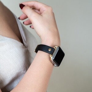 Op de afbeelding: Een zwart leren horlogebandje met een gouden gesp en een zilveren Apple Watch.