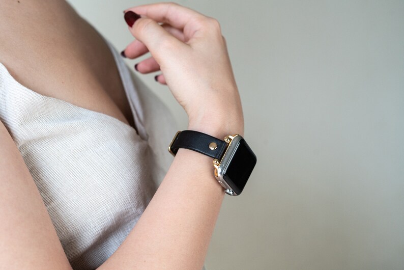 Op de afbeelding: Een zwart leren horlogebandje met een gouden gesp en een zilveren Apple Watch.