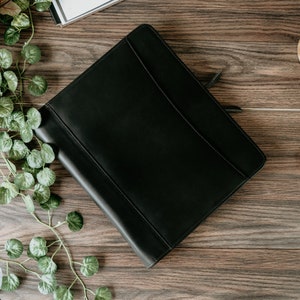 May include: A black leather bound journal with a zipper closure. The journal is lying on a wooden surface.