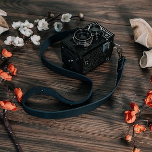 May include: A black vintage camera with a blue leather strap, surrounded by white and orange flowers on a wooden surface.