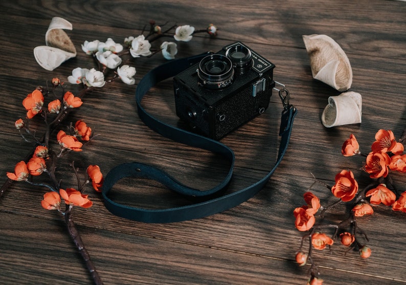 May include: A black vintage camera with a blue leather strap, surrounded by white and orange flowers on a wooden surface.