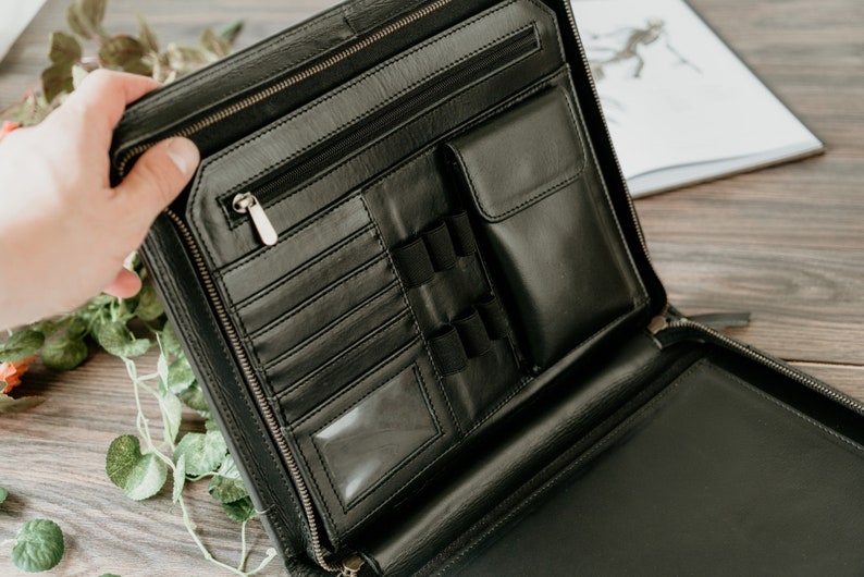 May include: Black leather zippered portfolio with multiple pockets and compartments. The portfolio is open and the interior is visible.