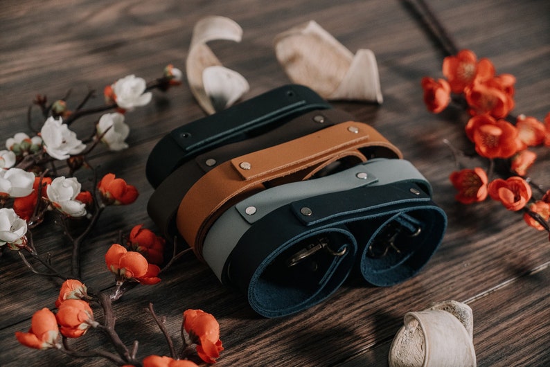 May include: Four leather camera straps in different colors: dark blue, light blue, brown, and black. The straps are stacked on top of each other and have metal hardware.