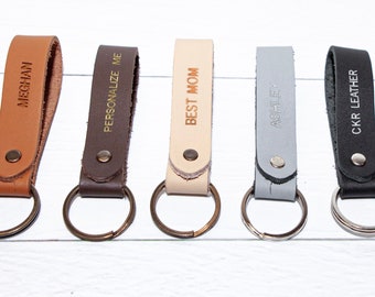 May include: Five leather keychains with metal rings. The keychains are brown, dark brown, beige, gray, and black. The keychains are personalized with the text "Meghan", "Personalize Me", "Best Mom", "Ashley", and "CKR Leather".
