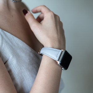 May include: A light gray leather watch band with a rose gold buckle and a silver Apple Watch.