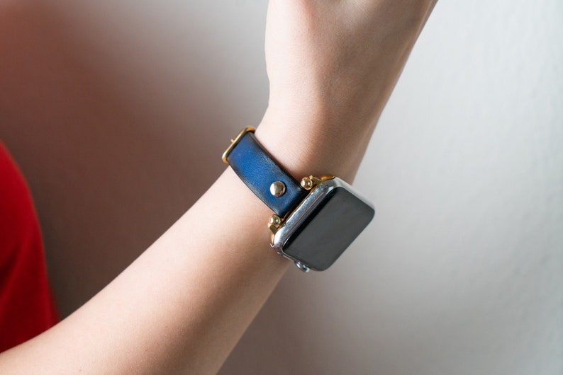 Op de afbeelding: Een zilveren Apple Watch met een donkerblauwe leren band. De band heeft gouden accenten en het horloge heeft een zwart display.