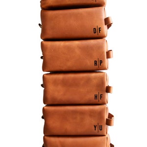 May include: A stack of six brown leather toiletry bags with personalized initials. The initials are E|C, D|F, R|P, H|F, Y|U, and K|L.