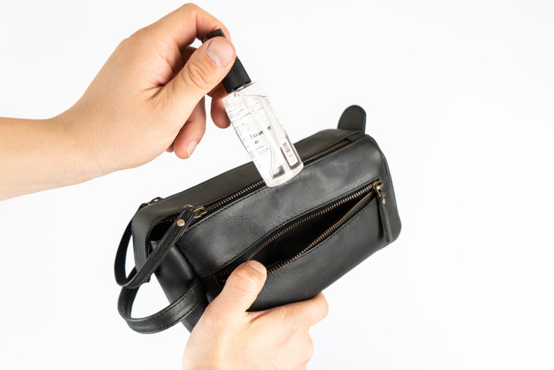 May include: A black leather travel bag with a zipped opening. A hand is holding a clear bottle of hand sanitiser and placing it inside the bag.