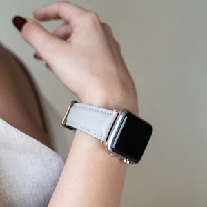 May include: A light gray leather watch band with a rose gold buckle and a silver Apple Watch.