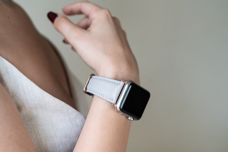 May include: A light gray leather watch band with a rose gold buckle and a silver Apple Watch.