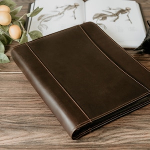 May include: A brown leather zippered binder on a wooden surface. The binder has a smooth, textured surface and is closed.