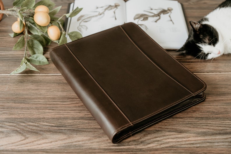 May include: A brown leather zippered binder on a wooden surface. The binder has a smooth, textured surface and is closed.