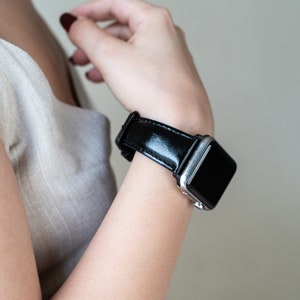 May include: A black leather watch band with a silver watch face. The band is shiny and has a smooth texture.