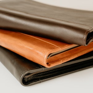 May include: Three leather zippered folders stacked on top of each other. The top folder is brown, the middle folder is orange, and the bottom folder is black.