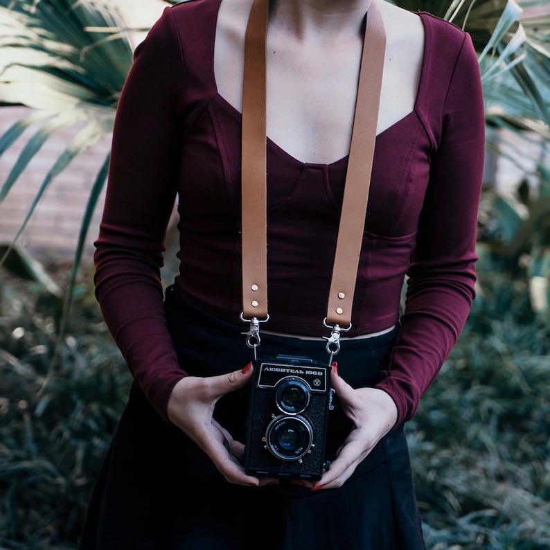 May include: A person wearing a maroon top and a black skirt is holding a black camera with a brown leather camera strap. The camera has a silver lens and the text "АВТОМАТ 1968" is printed on the front.