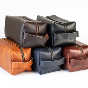 Op de afbeelding: Vijf leren reistassen in verschillende kleuren, op elkaar gestapeld. De tassen zijn bruin, beige, donkerblauw en bordeauxrood.